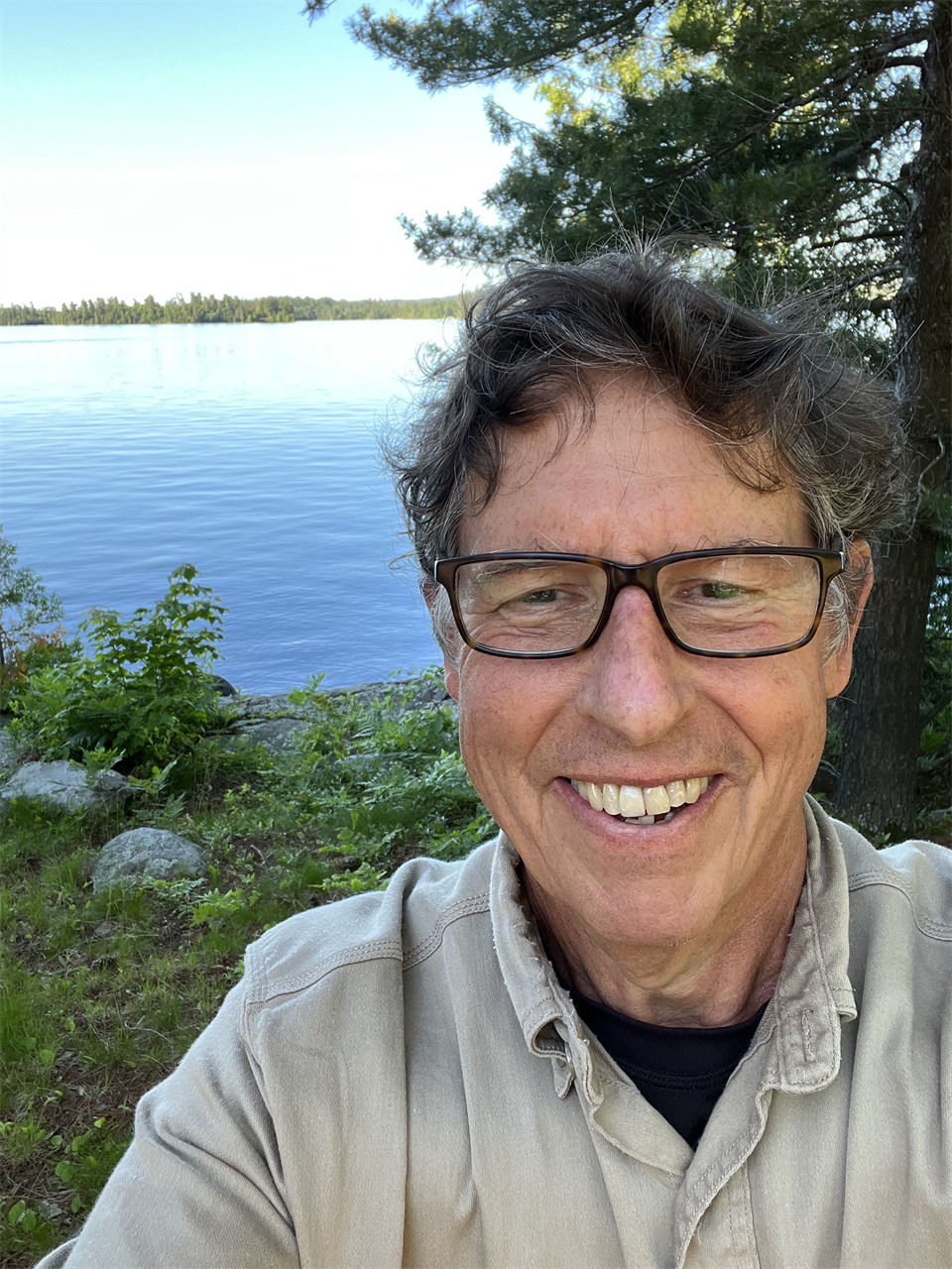 Middle-aged caucasian presenting man wearing glasses smiling standing outdoors in front of a lake with conifer tree and grass in the background.