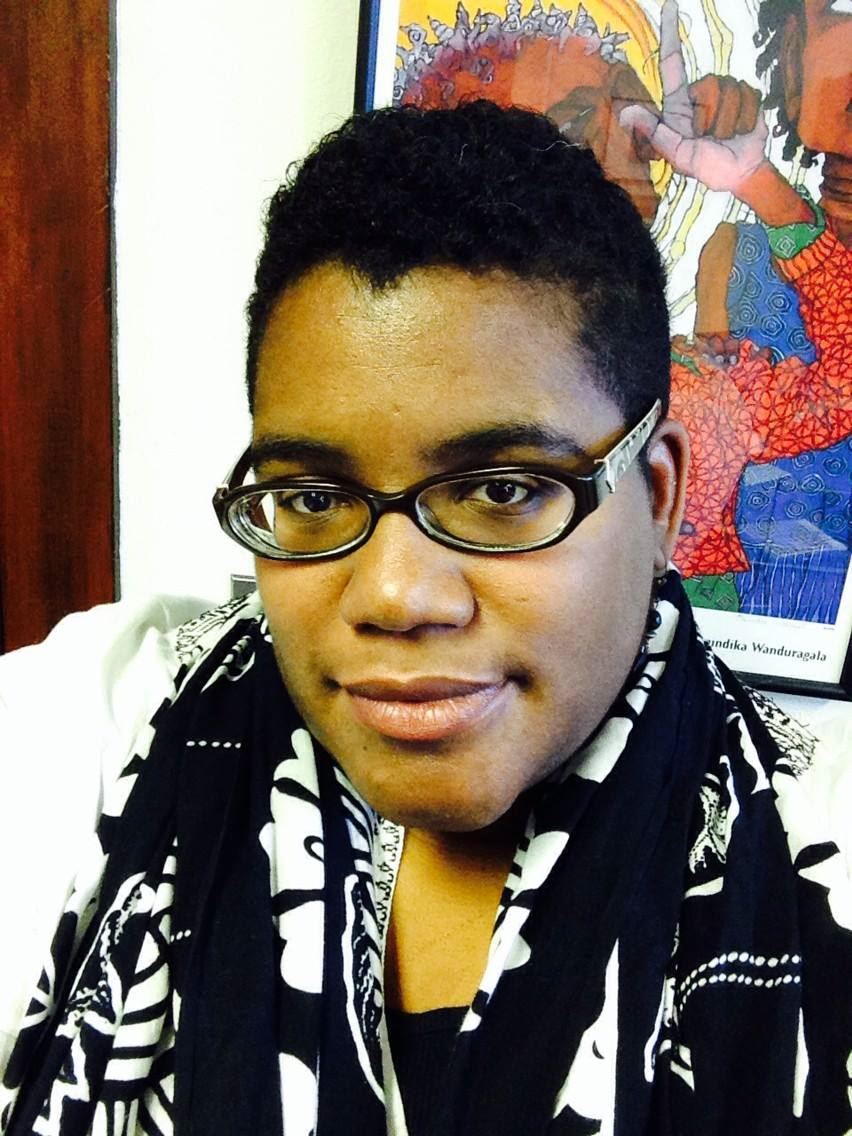 Headshot of a Black woman with short hair and glasses wearing a black and white graphic scarf with colorful artwork on the wall behind her.